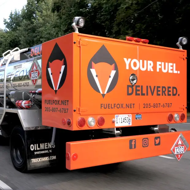 The back of an orange FuelFox tanker truck, branded "Your Fuel. Delivered," driving down a tree-lined road.